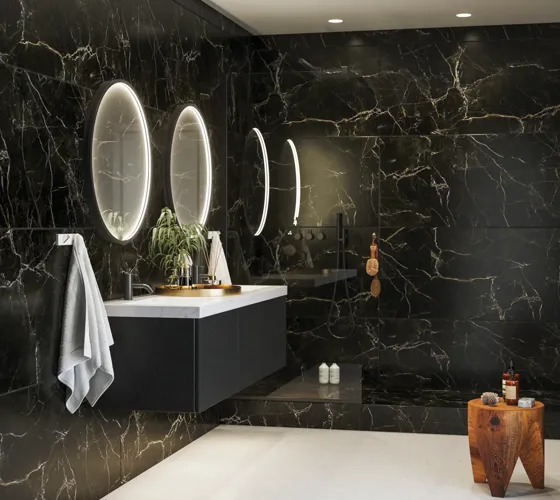 Elegant and modern bathroom inspiration with dark marble walls, round illuminated mirrors with black frames, wall-mounted vanity unit with double sinks, and a spacious shower area